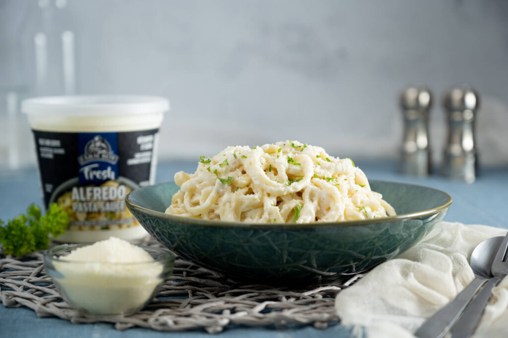 Bowl of fresh pasta with alfredo sauce on a table with farm Boy Alfredo Sauce, small bowl of parmesan cheese, and salt and pepper shakers