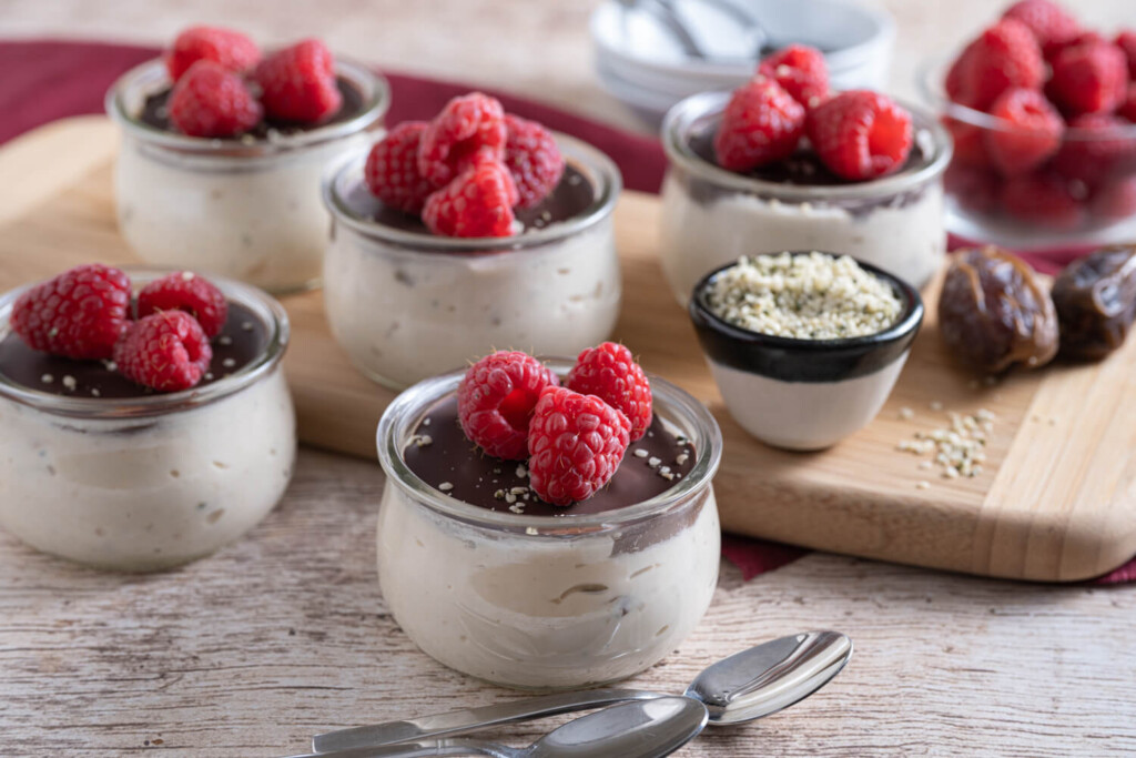 Small glass jars of yogurt, nut, and date pudding with chocolate and fresh raspberries on top for a healthy dessert recipe
