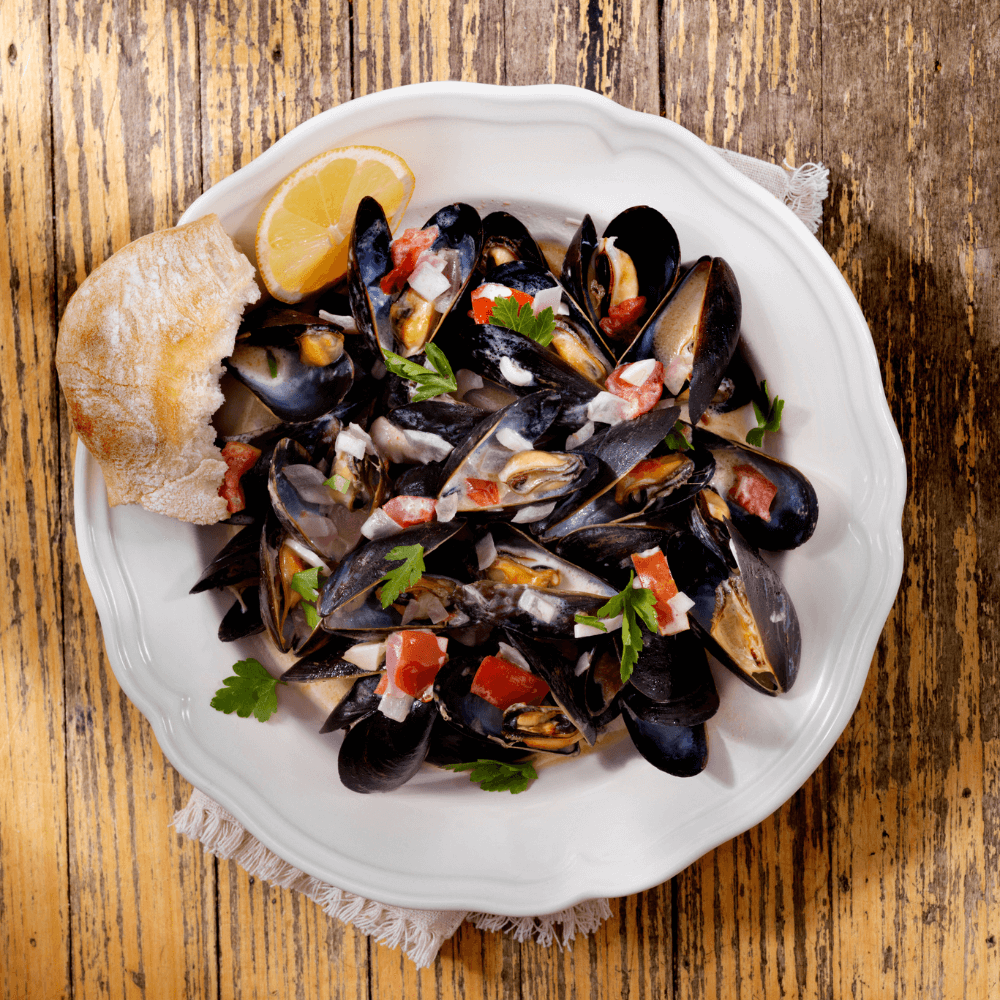 White plate of mussels with garlic, tomatoes, peppers, and herbs on a wooden table with a wedge of lemon and piece of crusty bread
