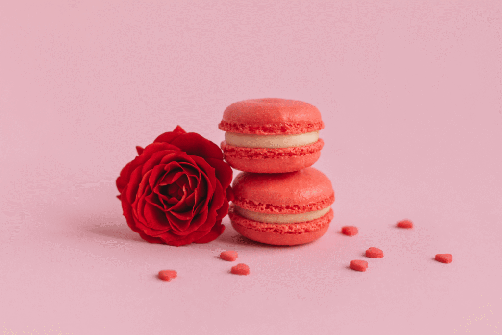 Tasty French macaroons with red rose on a pink pastel background, decorated with sweet hearts
