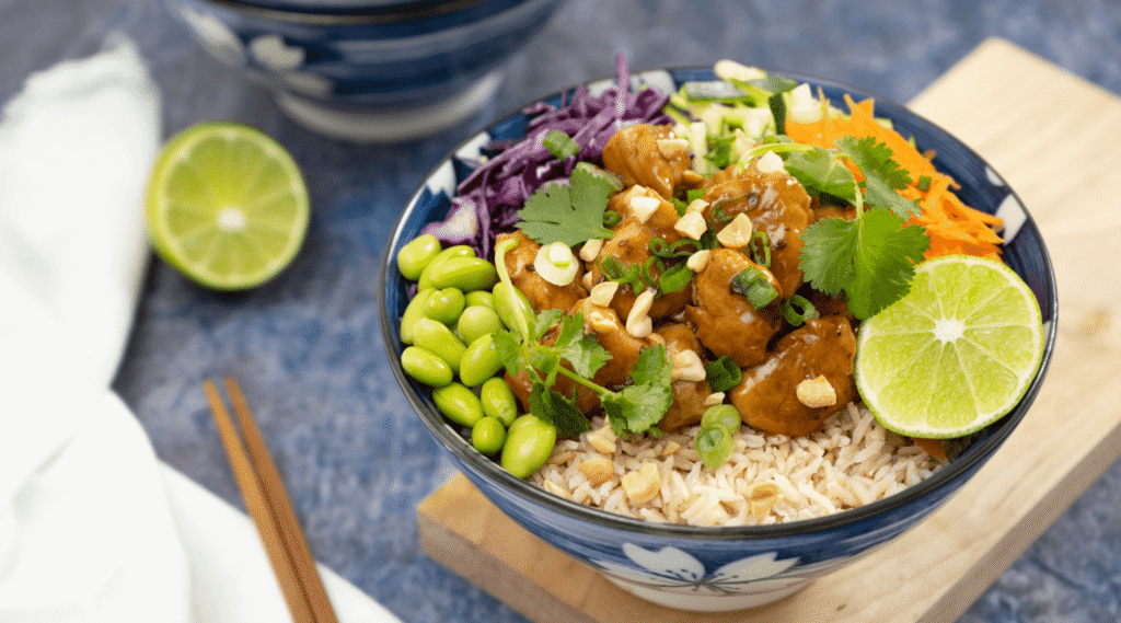 Vegan dinner: Blue bowl filled with rice, vegan plant-based chicken, edamame, red cabbage, and carrots, garnished with chopped peanuts, cilantro, and half a lime.