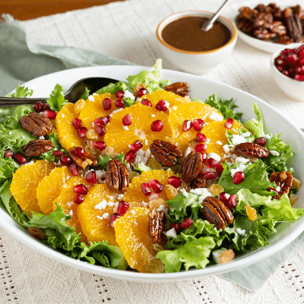 Salad on plate. Salad has mixed greens, sliced oranges, candied pecans, crumbled feta cheese, and pomegranate arils