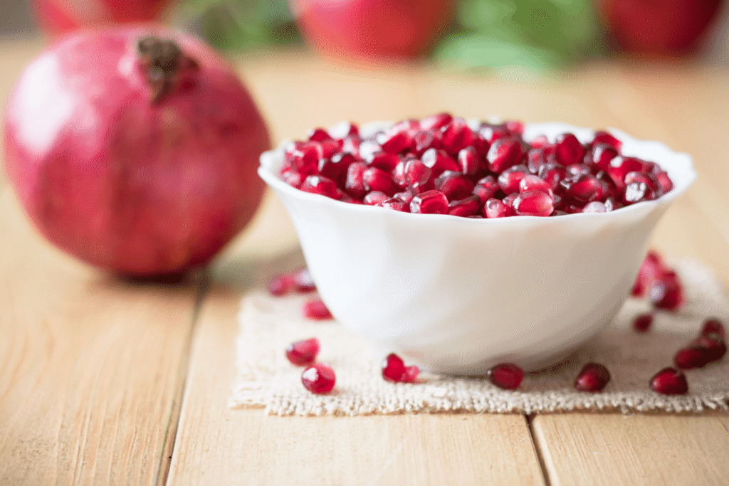 Small bowl of pomegranate arils on wooden table with pomegranate in background