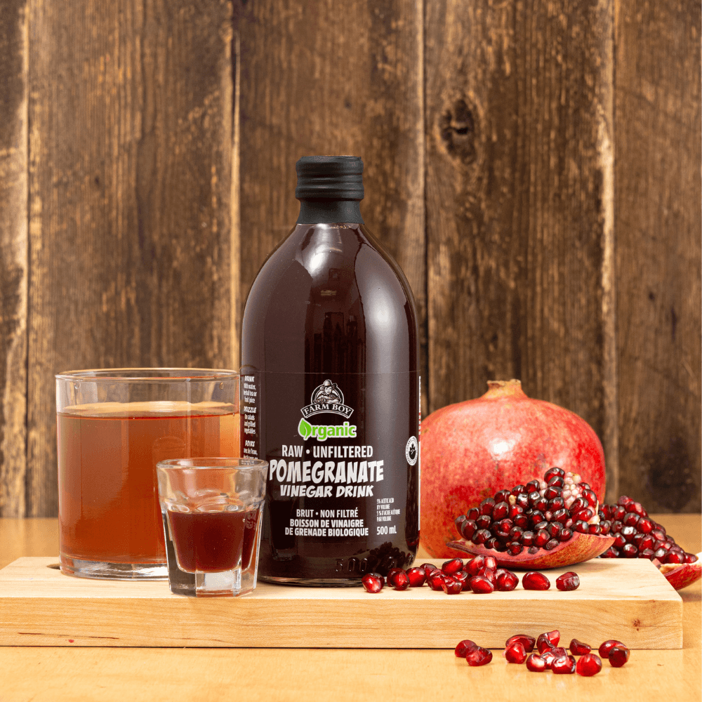 image of bottle of Farm Boy Pomegranate Vinegar Drink with glass of drink, shot glass, whole pomegranate, and segment of pomegranate with arils spilling out