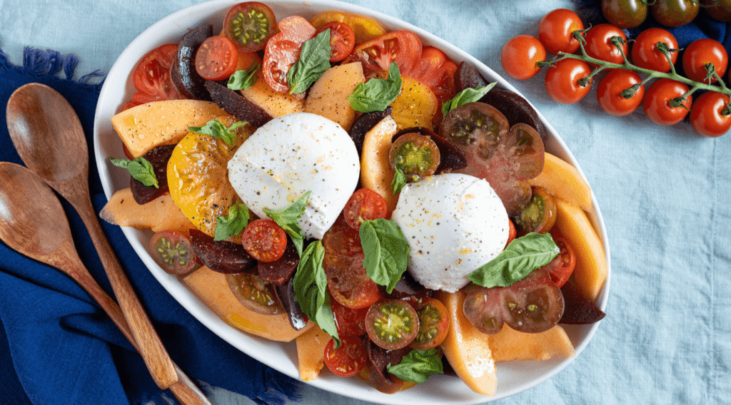Overhead image of platter of heirloom tomato, beet, and cantaloupe salad finished with balls of fresh mozzarella and torn basil leaves