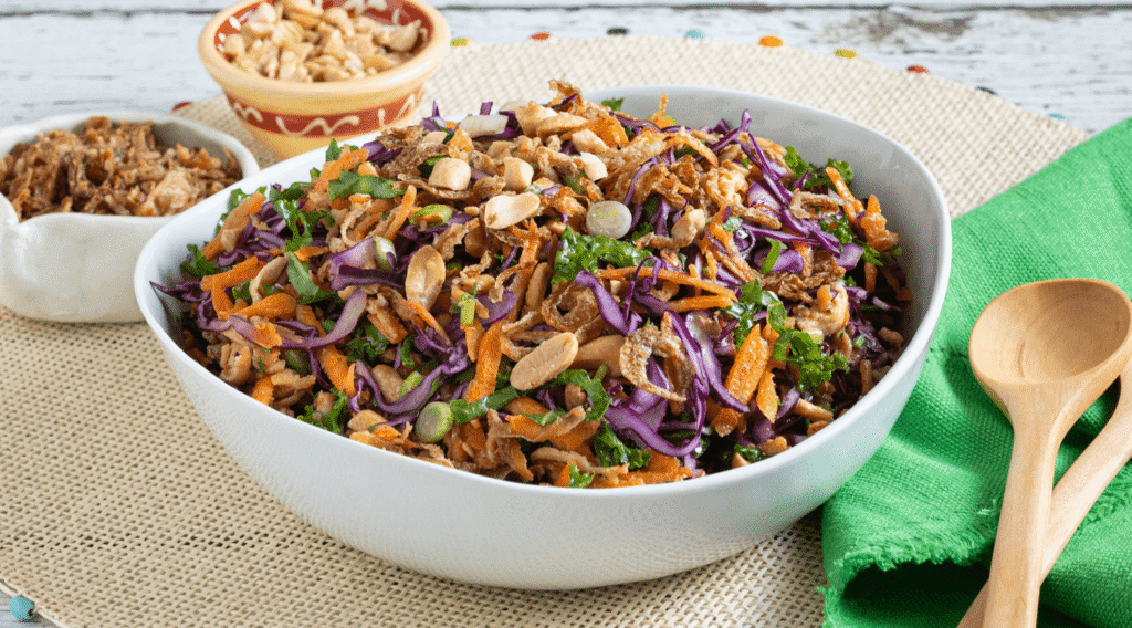 Woven placemat with white bowl of rainbow tahini tofu slaw with shredded carrots, kale, cabbage, tofu, peanuts, and crispy onions