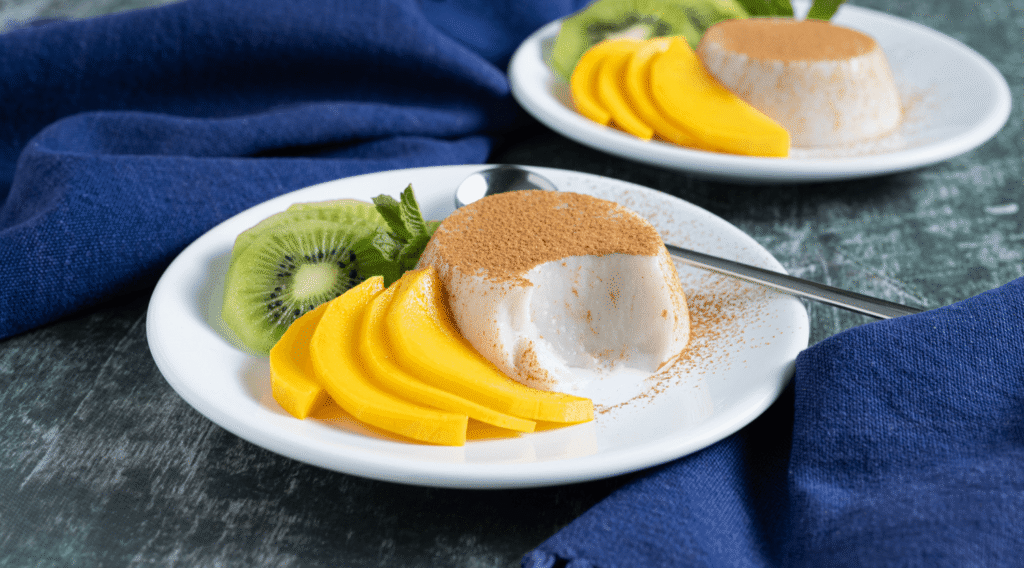 White plate with individual portion of spiced coconut pudding, dusted with cinnamon and garnished with thinly sliced kiwi and mango