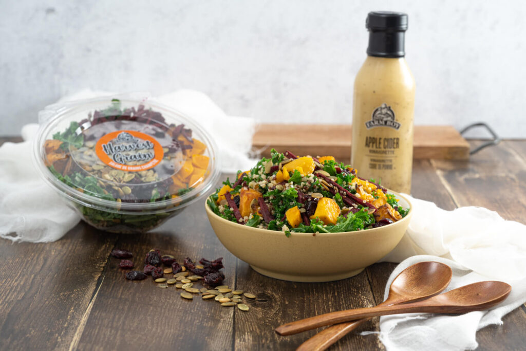 Plated bowl of Farm Boy Harvest Grain Salad with wooden serving spoons and bottle of Farm Boy Apple Cider Vinaigrette in background
