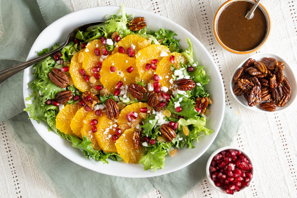 Overhead image of serving dish with salad made with orange slices, pomegranate arils, greens, cheese, and candied pecans