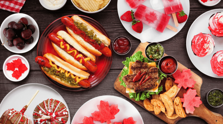 Canada Day patriotic food. Above view table scene on a dark wood background. Hot dogs, hamburgers and maple leaf red and white theme snacks.