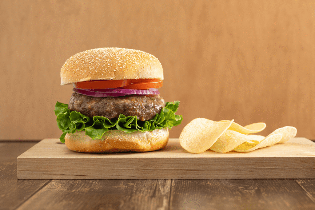 Farm Boy Gourmet Burger topped with lettuce, tomato, and red onion on cutting board with potato chips
