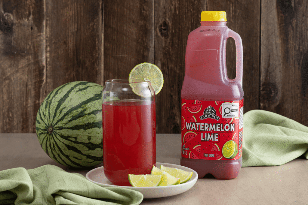 Glass of red juice with bottle of Farm Boy Watermelon Lime Fruit Drink beside it, accompanied by whole watermelon and sliced lime wedges