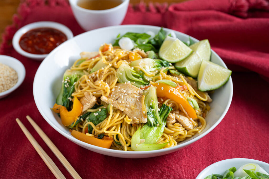 White bowl of chicken lo mein with fresh veggies and sesame seeds