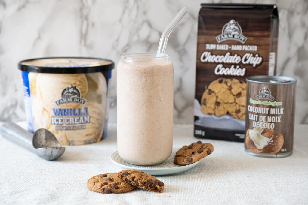 Talle glass of chocolate coconut vailla milkshake with glass straw, flanked by package of chocolate chip cookies, can of coconut milk, and tub of Farm Boy Vanilla Ice Cream