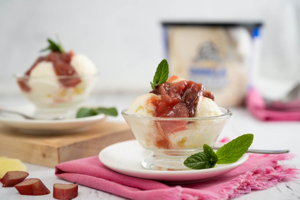 Small glass bowl with two scoops vanilla ice cream with roasted rhubarb topping and fresh mint leaves