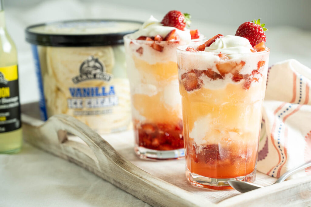 Two tall glasses with layers of vanilla ice cream, chopped strawberries, whipped cream, and sparkling lemon lime soda