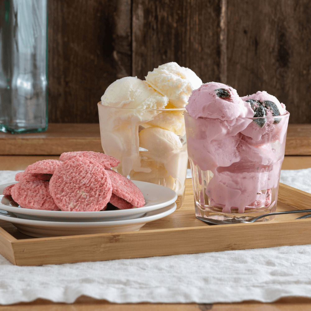 Two glasses with multiple scoops of different ice cream beside plate of pink cookies