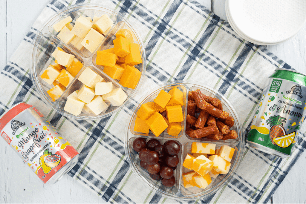 Overhead image of two plastic segmented snack boxes of cheese, grapes, and sausages on checked cloth