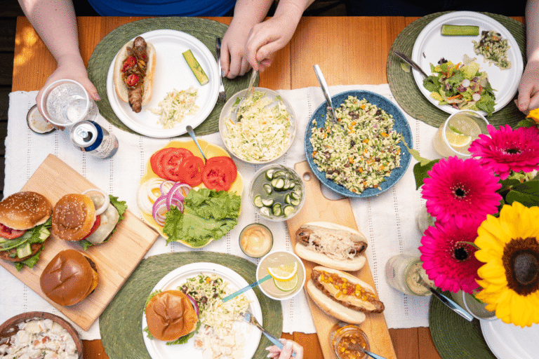 Overhead image of end-of-summer table with assorted bowls of salads, gourmet burgers, artisan sausages, fresh flowers, and drinks