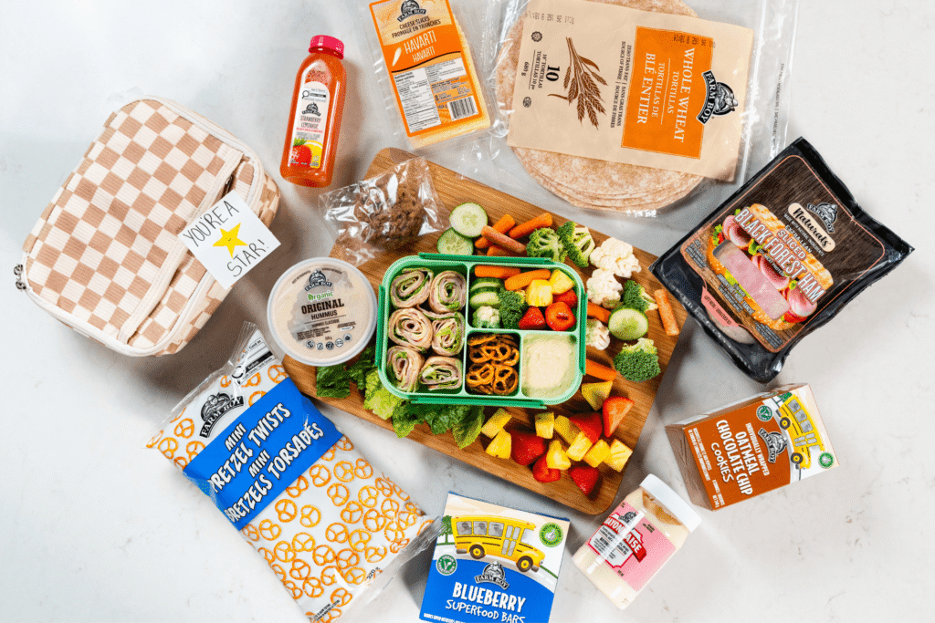 Top view of lunch bento box with wrap roll-ups, fresh veggies, dip, and pretzels, surrounded by assorted Farm Boy items and a lunchbag